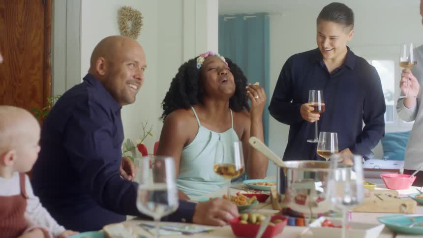 A group of people happy engage in conversation while sitting and standing around a dinner table in a house during the day