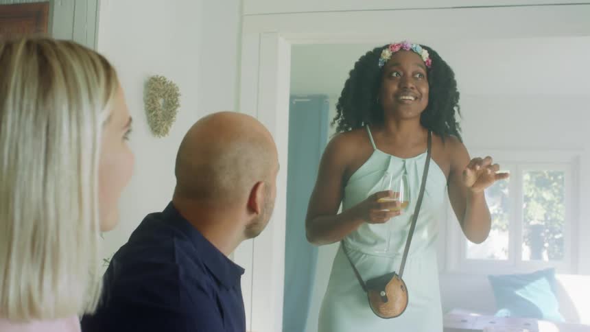 A woman standing in a house holds a glass of wine and talks to a man and woman
