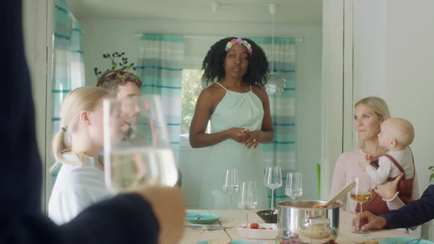 A woman stands at the end of a dining tables talking to friends seated at the table for a meal