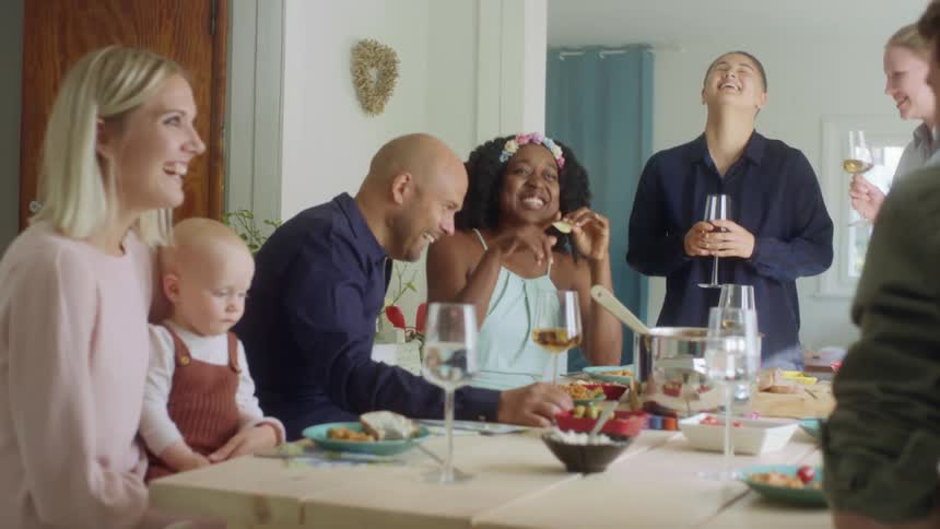 A group of people sit around a home dinner table with wine glasses and talking happily together