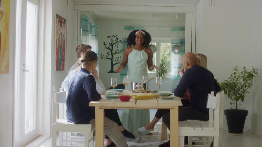 A woman stands in front of a home dinner table with other people sitting and listening to her talk