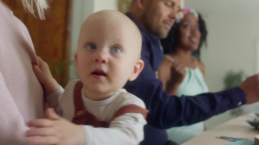 A baby is held in his or her mothers lap at a table with others eating at the table in the background