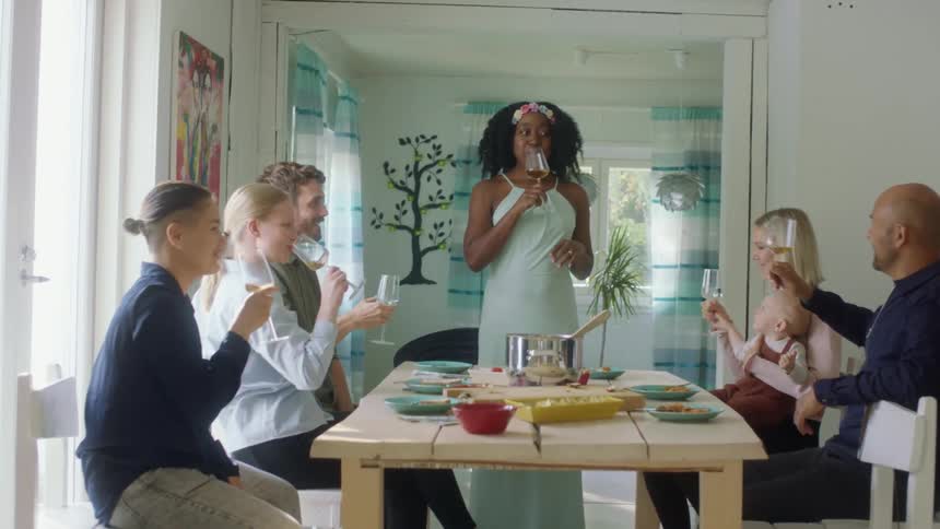 A woman standing at the head of a dining table in a house leads a toast before everyone sitting at the table takes a drink