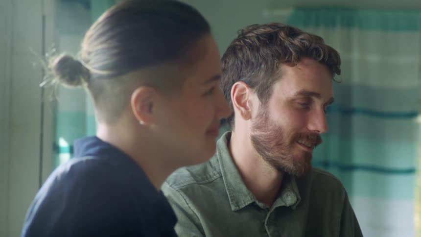 A man drinks a cup of coffee and laughs as he sits next to a woman at a table in a house