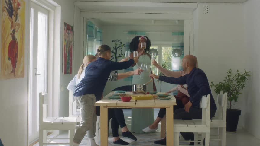 A woman stands at the head of a home dinner table giving a toast and everything taps their wine glasses together