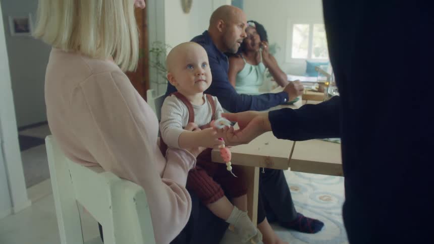 A group of friends relax around a dining table after eating and a mom holds her child in her lap as a friend retrieves a dropped toy