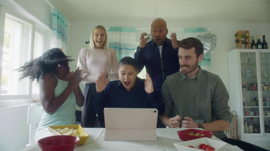 A group of people in a home sit and stand around a table and look at a computer and then cheer and high five one another