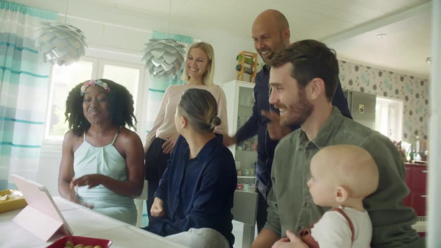A group of people cheer while watching a tablet on its stand on a table in a house during the day
