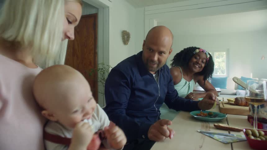 A bald man sitting at a home dining table talks with a baby being held in the lap of the woman sitting next to him