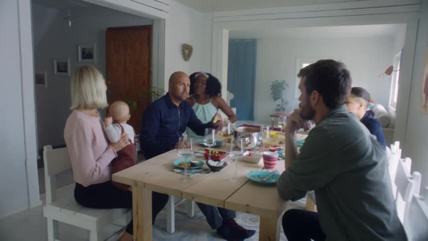 A woman carries a baby to a dinner table with other men and women sitting there
