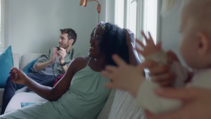 Friends relaxing on couches in a living room clap and snap fingers as a woman holds her baby