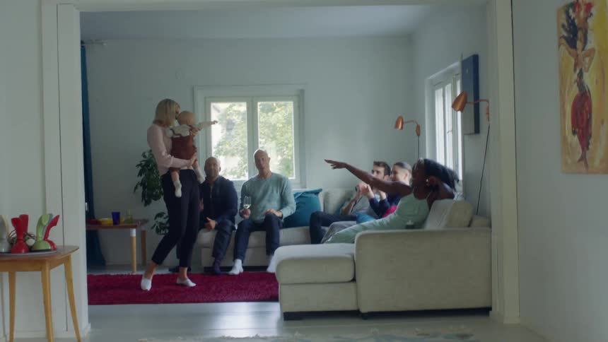 A group of people sit on a the couch dancing along to a woman standing in front of them dancing while holding a baby