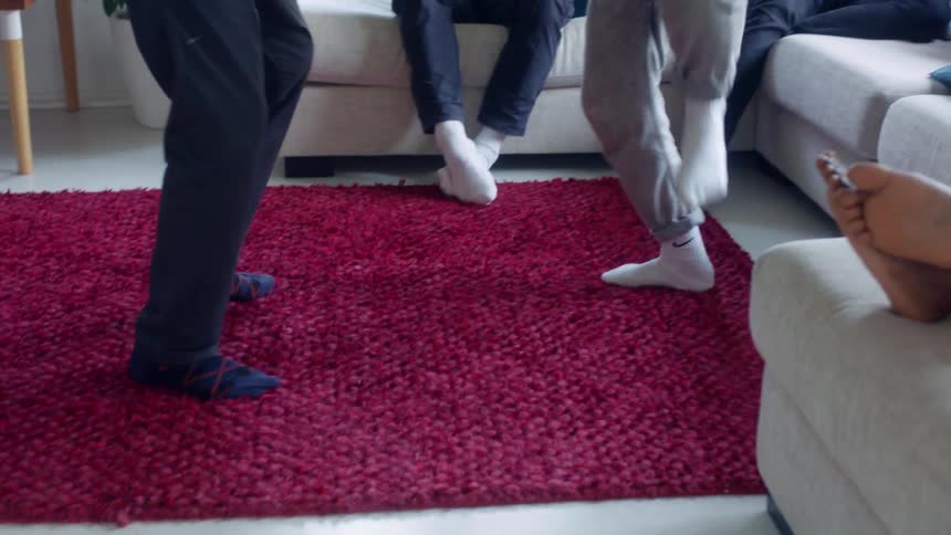 Two people are standing and dancing in front of other people sitting and lounging on a couch indoors