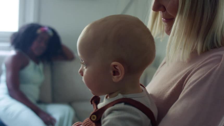 A mother stands and holds her child while a friend sits on a couch and watches