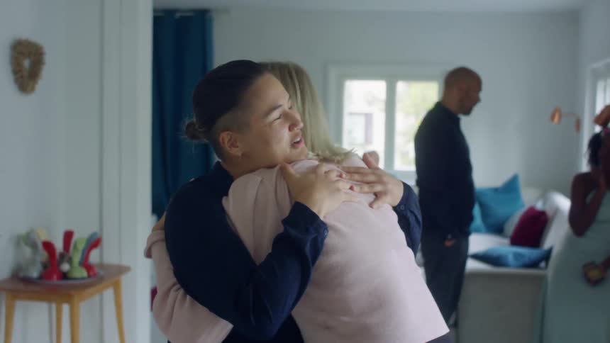 Two friends smile and hug in a house as other people stand behind them