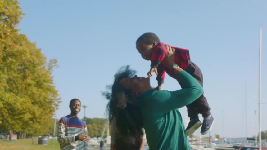 Mother playfully raises son from the ground while in a park near boats on a lake with her family
