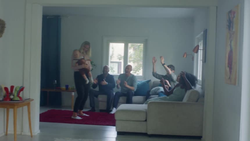 A woman dances with her baby as her friends sit on a couch and clap along