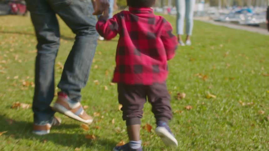 A teenager holds his toddler brothers hand as they run toward their parents in a park along the water