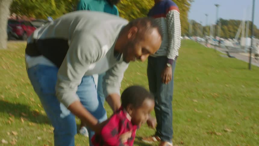 Father playfully lifts his infant son in the air over the green grass in a park