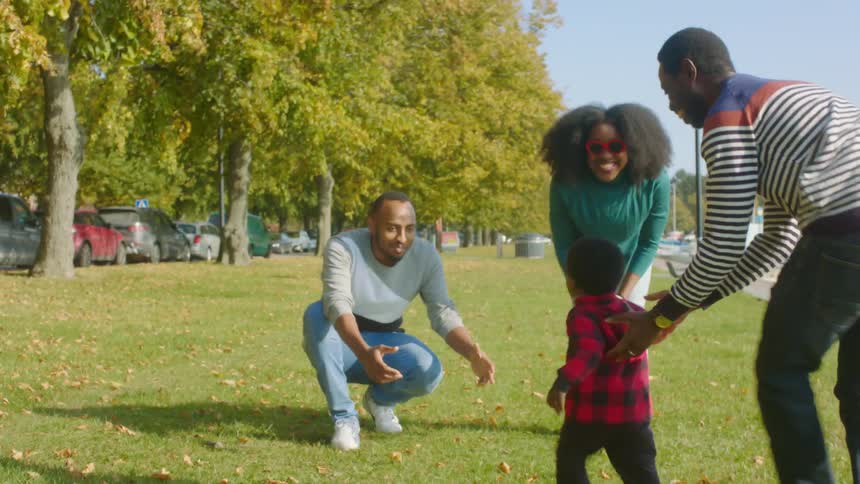 A man in a public park off the water runs with his little boy towards a man and woman and the man picks him up