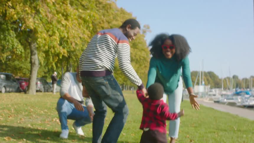 A man holds his toddlers hand and they run to his mom who lifts him up in the air