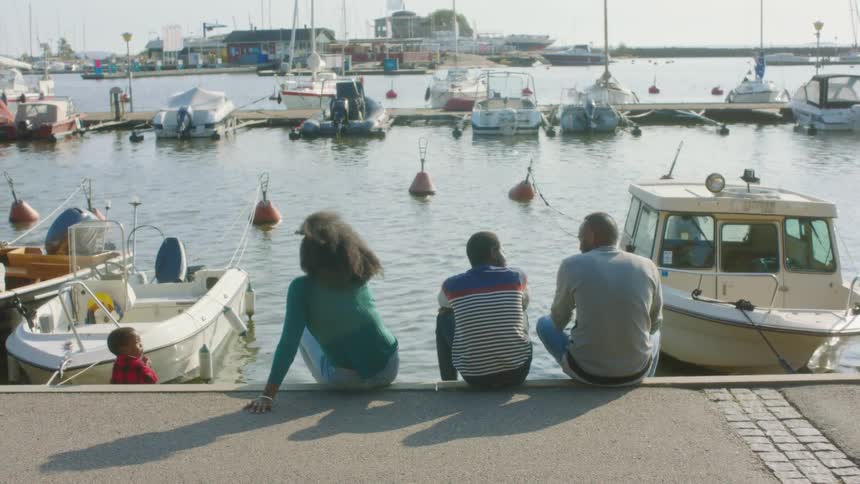 A mom and dad sit with their sons on a pier looking at boats and water during the day