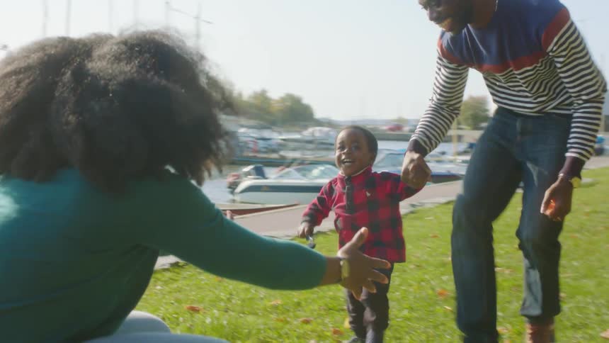 A man holds a little boys hand as they run over to a woman and they embrace next to a body of water with boats