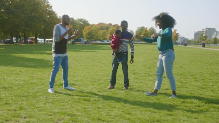 A man holds a small toddler in a large grass park and dances with another man and woman