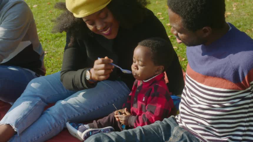 A mom feeds her son baby food as she sits with a man on the grass relaxing during the day