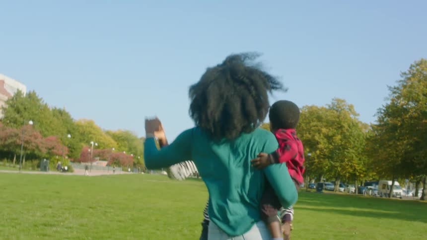 A woman holds her toddler son as she spins and dances in the park with her husband