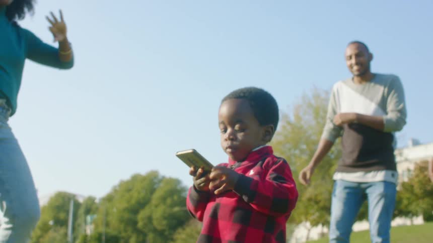 A toddler boy holds a cell phone while him and other adults behind him dance in an outdoor space