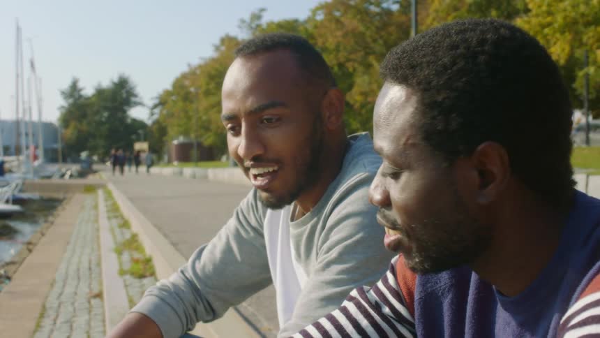 Two men sit talking near docked boats in water side park as people walk past
