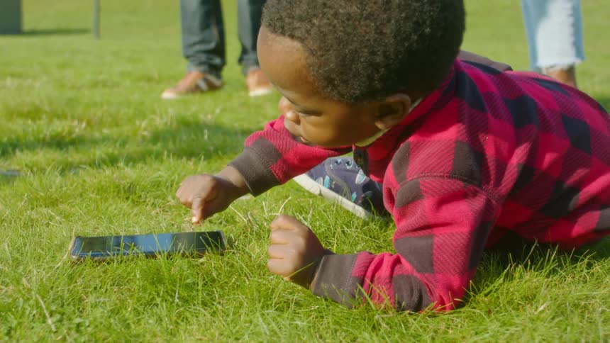 A young boy plays with a cell phone in the grass as his parents dance behind him