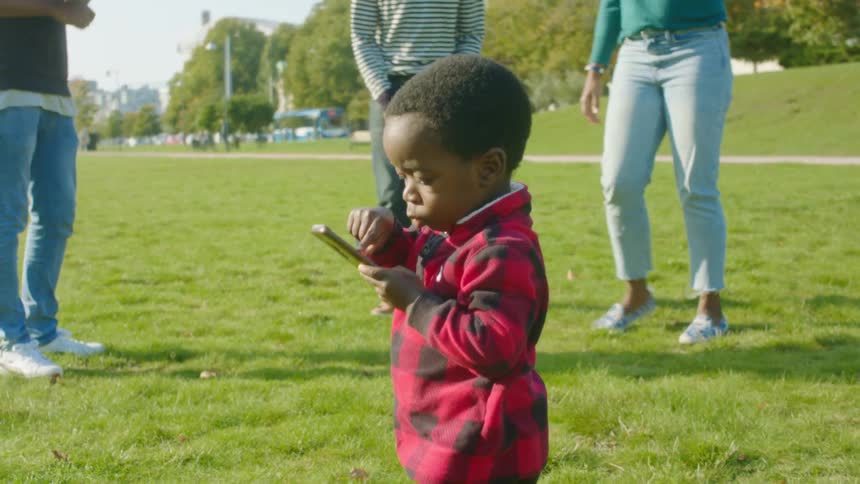 A toddler boy picks up a cell phone from the public park grass and starts to push buttons