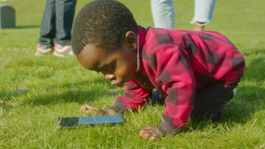 A little boy on the grass looks at a cell phone as his mom and dad dance behind him