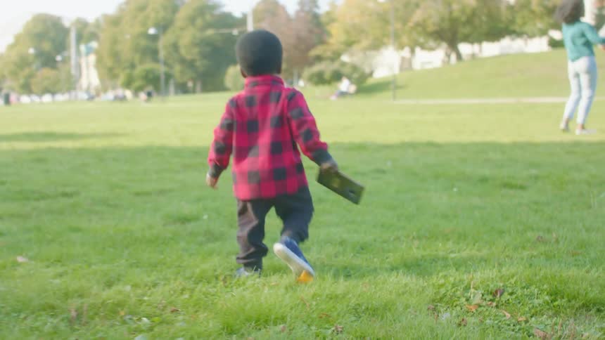 A little black boy walks on the grass holding a cell phone as he heads towrds a group of three adults