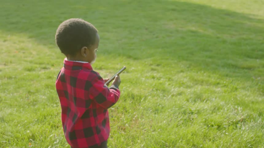 A little boy standing on a grassy field holding and playing with a smart phone