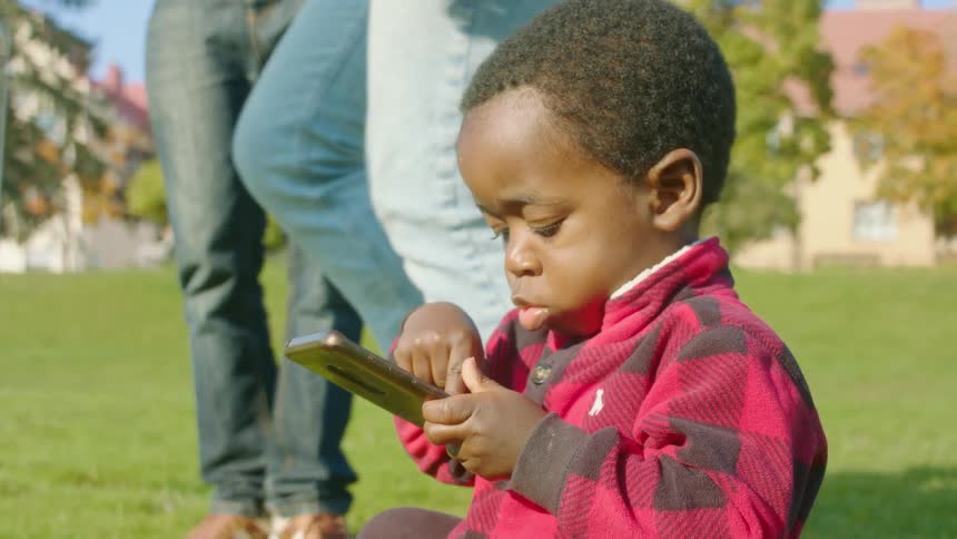 A young boy plays on a smartphone in the park while his parents dance above him