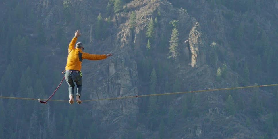 A person sly stands up on a tight rope bridging a mountain