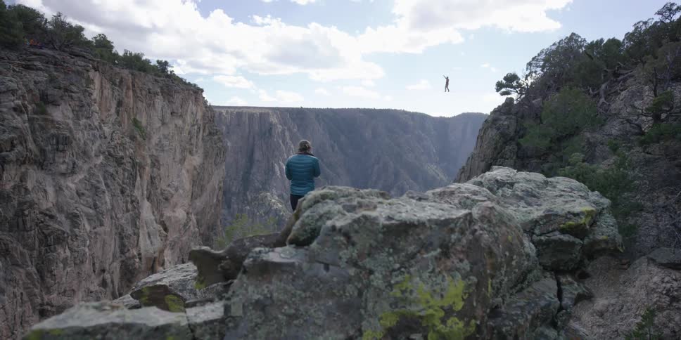 A woman stands on a precipice and watches someone walking a rope over a gorge