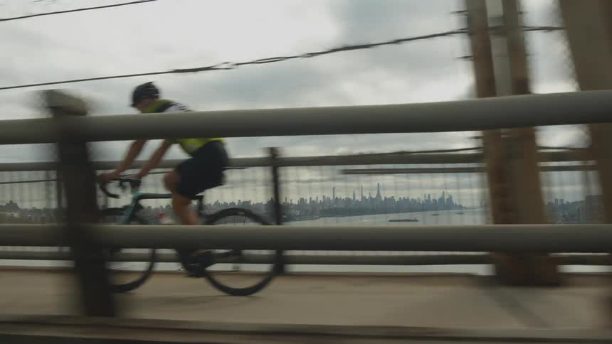 Bicyclists riding over a large city bridge with skyscrapers in the distance