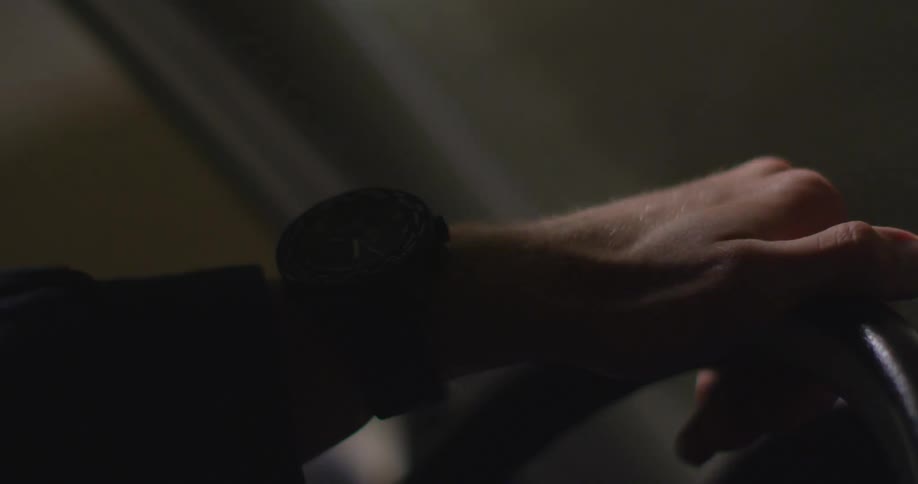 A males hand is on a moving vehicles steering wheel as it drives at night