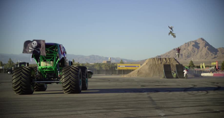 Multiple people drive race cars and jump large dirt hills on dirt bikes with a big monster truck sitting in the area