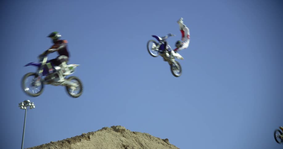Four people jump dirt hill on dirt bikes and do stunts on a sunny day with a light in the background