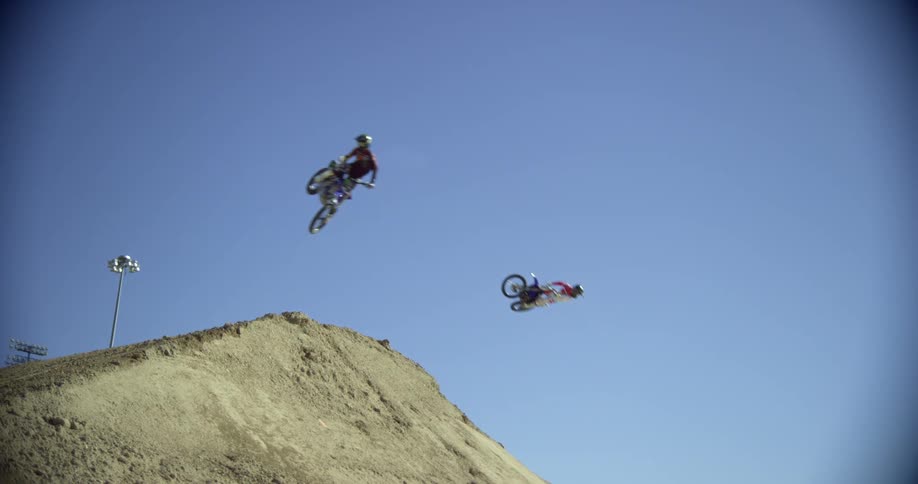 A group of motocross riders fly through the air over a mound of dirt on their dirt bikes one after another under a clear daytime sky