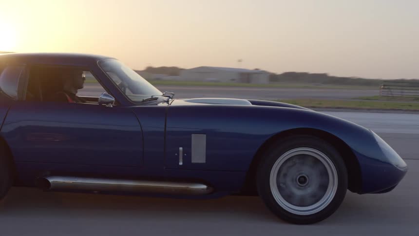 A blue sports car drives on a runway at an airport at dusk
