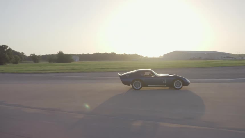 A man in a vintage sports car speeds through a parking lot next to a field