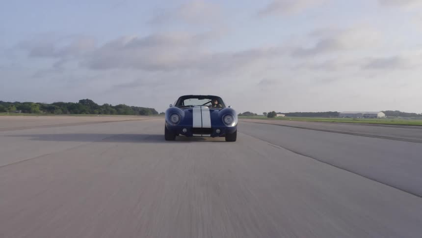 A man drives a blue vintage coupe with white racing stripes down a straight track