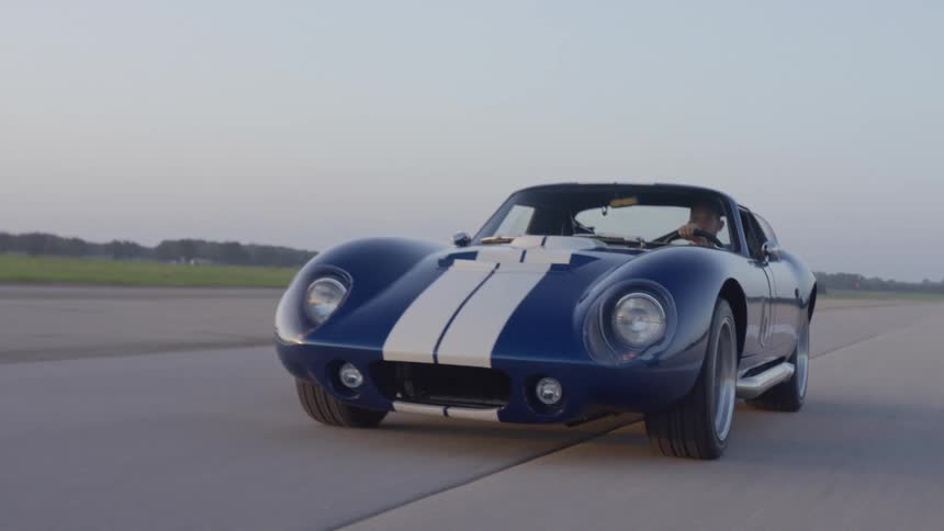 A man drives a sports car down a runway in a flat field during the day