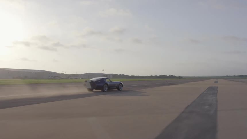 A sports car drives fast along an empty race track in a rural setting